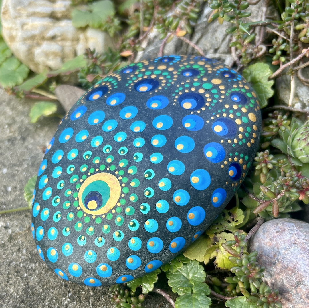 Stein mit einem Mandala bemalt in Farbtönen einer Pfauenfeder im Garten liegend