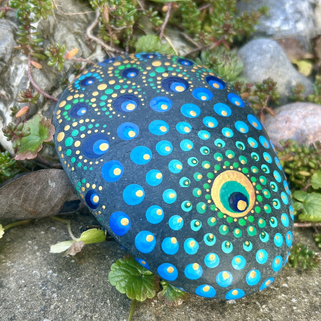 Stein mit einem Mandala bemalt in Farbtönen einer Pfauenfeder im Garten liegend