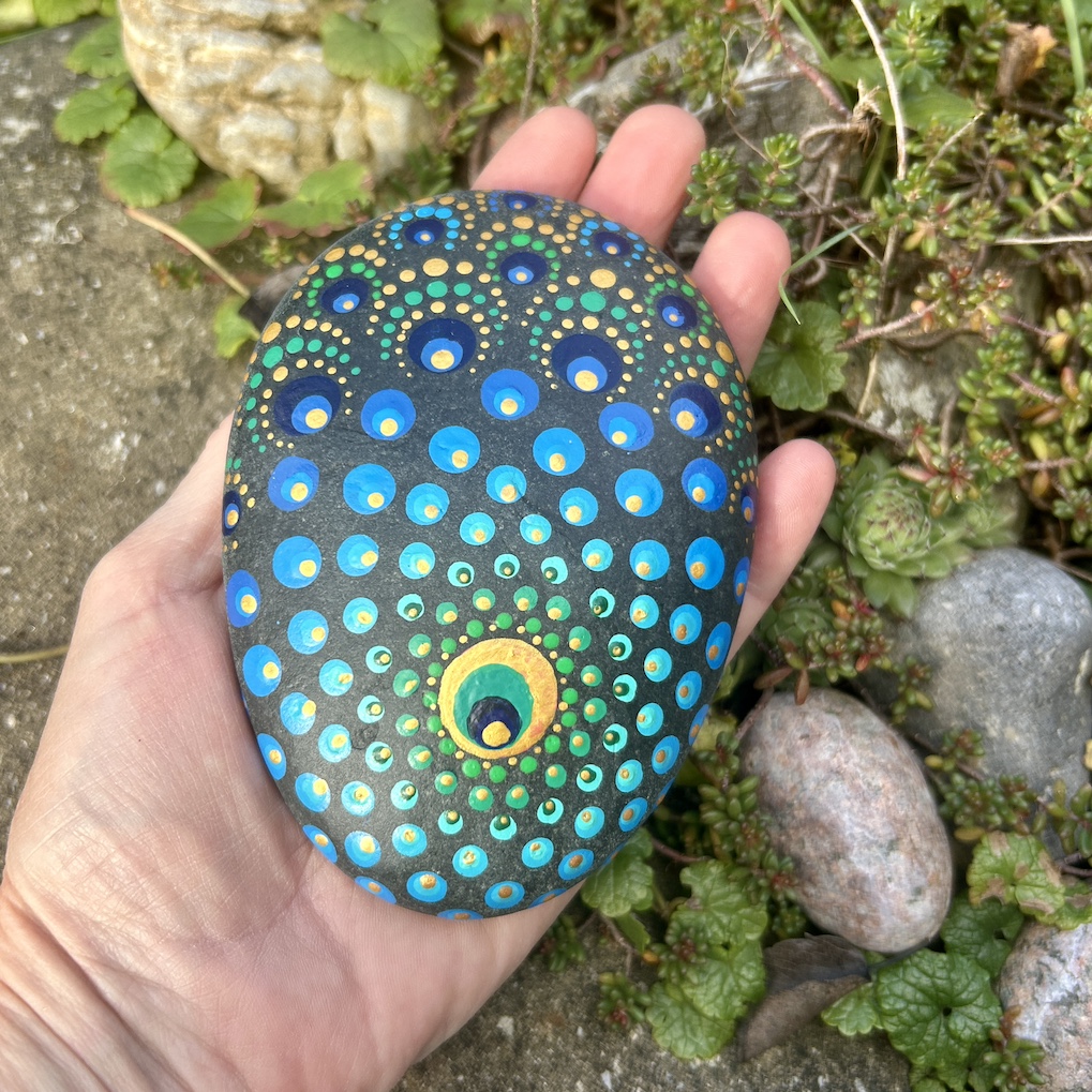 Stein mit einem Mandala bemalt in Farbtönen einer Pfauenfeder in der Hand gehalten